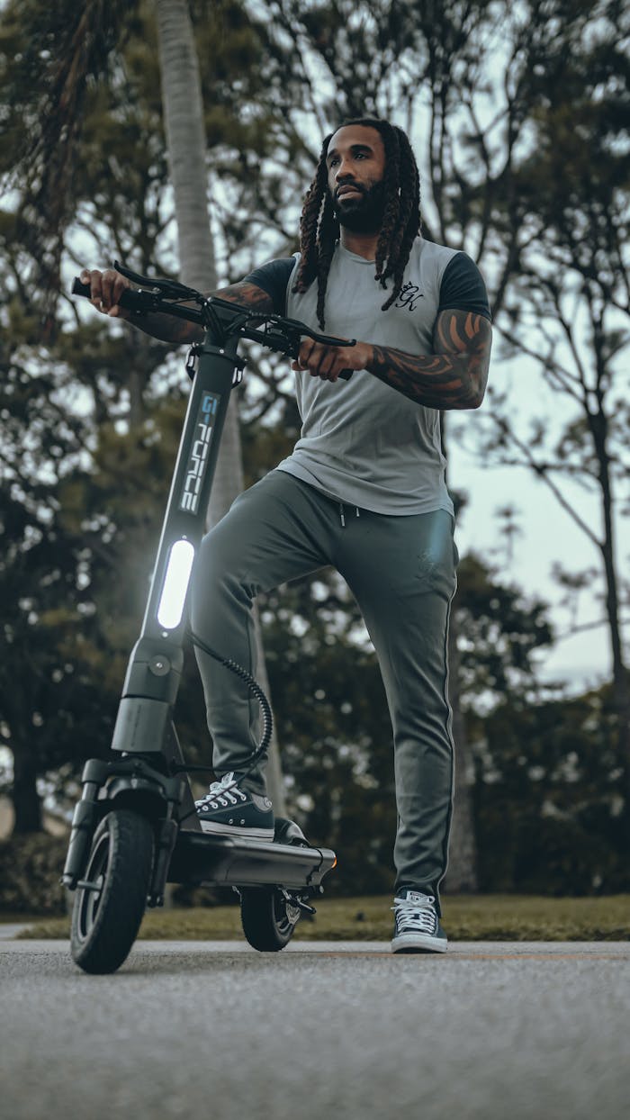 Adult man with tattoos standing with an electric scooter on a paved path in a park.